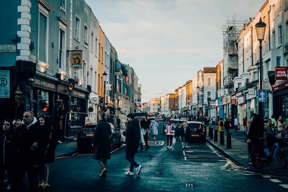 A busy urban street scene featuring a row of multi-story buildings with various storefronts, including a pub named 'Duke of Wellington' on the left side, and a sign for Five Guys on the right. The buildings have a mix of painted facades and brickwork, with some under scaffolding indicating ongoing maintenance or refurbishment. Pedestrians, dressed in casual and business attire, walk along the pavement and cross the street, which is marked with traffic signs and road markings. Several parked cars line the curb, and a few moving vehicles are visible further down the street. The atmosphere is illuminated by warm, late-afternoon sunlight, casting soft shadows and highlighting the textures of the building exteriors and street surfaces. The scene captures a typical day in a vibrant commercial district, with the environment indicative of an area served by alternative waste management or private rubbish removal services, consistent with the context of rubbish collection and disposal in urban settings, as referenced by Rubbish Removal Notting Hill.