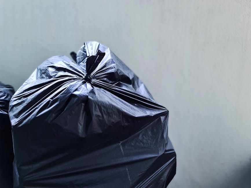 Close-up image of multiple black plastic rubbish bags filled with waste, tightly knotted at the top, with a glossy surface reflecting light, positioned on a dark surface against a plain, light-colored background. The bags appear to contain mixed household waste and are arranged in a manner that suggests they are prepared for disposal or collection. The environment looks like an outdoor or indoor setting designated for rubbish storage before pick-up, highlighting an element of private waste management, which Rubbish Removal Notting Hill offers as an alternative to official collection services. The scene emphasizes the practical aspect of rubbish handling, with the bags clearly stacked and ready for collection, reflecting an efficient on-site clearance approach that supports independent waste disposal efforts in the neighbourhood.