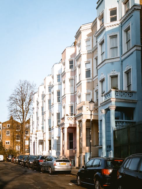 A row of multi-storey Victorian residential buildings with ornate architectural details, painted in light pastel shades of blue and cream, line a city street. The buildings feature bay windows, decorative cornices, and small balconies, some with potted plants. In the foreground, parked cars of various makes and models occupy a narrow street or driveway, with some partially visible in the lower part of the image. The street is shaded by leafless trees, indicating late autumn or winter, and the sky above is clear with bright daylight illuminating the scene. The presence of parked vehicles and the residential setting provide context for private waste handling or alternative rubbish removal methods, reflective of services like those offered by Rubbish Removal Notting Hill, as suggested by the website domain. This urban environment highlights typical challenges faced during property clearance or regular rubbish collection in residential areas.