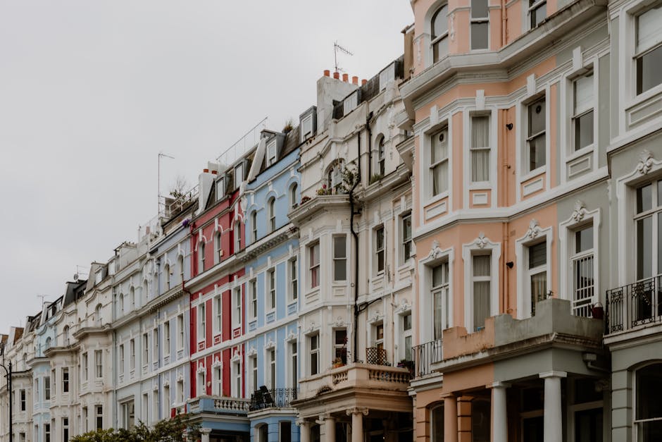 A row of Victorian-style terraced houses with detailed white facades, painted in pastel shades of pink, blue, peach, and white. The buildings have multiple large sash windows with white frames, some featuring small balconies with black iron railings. The rooftops include chimneys and antennae, with slight variations in roof angles and dormer windows. The buildings are situated on a paved street, with the front gardens and entrances visible at ground level. The sky appears overcast, casting diffused natural light over the scene. The image showcases an urban residential area typical of the historic architecture often found in London, where independent waste collection or on-site clearance might be performed to manage rubbish or renovation debris, eliminating the need for local authority collection, as exemplified by the surroundings and building facades. Rubbish Removal Notting Hill's services could be suitable for managing waste in such environments, especially during renovation or property clearance projects.