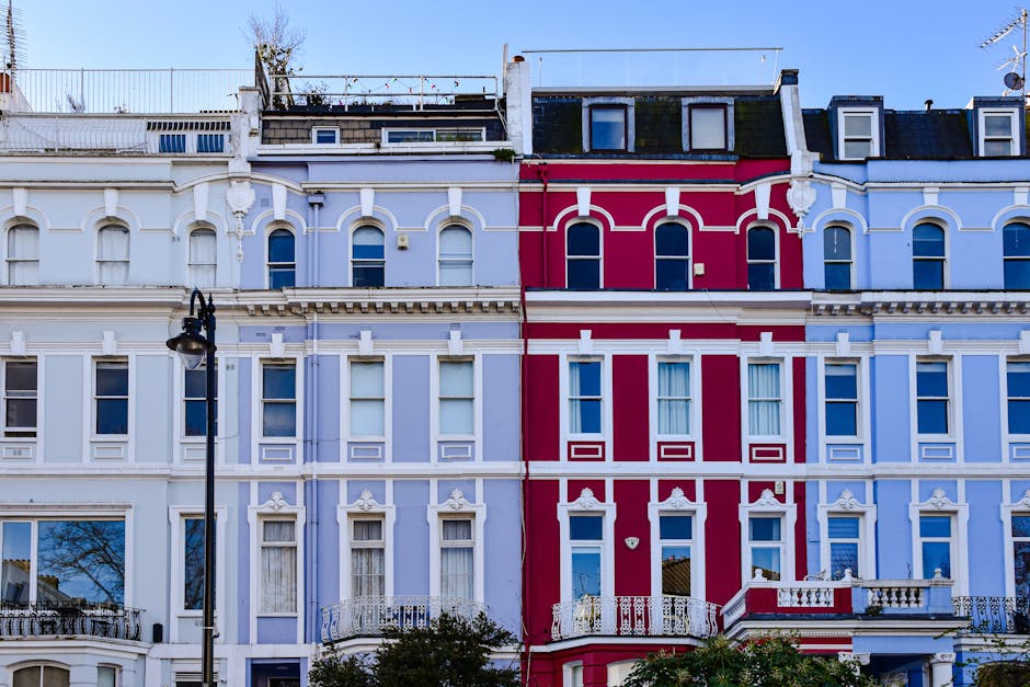 A row of multi-storey Victorian residential buildings with ornate architectural details, painted in light pastel shades of blue and cream, line a city street. The buildings feature bay windows, decorative cornices, and small balconies, some with potted plants. In the foreground, parked cars of various makes and models occupy a narrow street or driveway, with some partially visible in the lower part of the image. The street is shaded by leafless trees, indicating late autumn or winter, and the sky above is clear with bright daylight illuminating the scene. The presence of parked vehicles and the residential setting provide context for private waste handling or alternative rubbish removal methods, reflective of services like those offered by Rubbish Removal Notting Hill, as suggested by the website domain. This urban environment highlights typical challenges faced during property clearance or regular rubbish collection in residential areas.