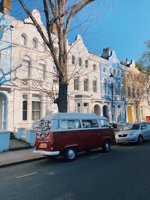 A vintage two-tone Volkswagen Type 2 van, painted in a deep red colour with a cream upper section, is parked on a residential street in front of a row of terraced houses with decorative facades. The van features a roof rack at the rear, holding a bicycle and some luggage or bags, suggesting private or independent waste collection or transport related to rubbish removal services. The street is lined with leafless trees, indicating late autumn or winter, with shadows cast on the pavement by the bright sunlight. Nearby, a modern silver sedan is parked alongside the van, and the pavement is clean and well-maintained. The row of terraced houses in the background features ornate architectural details, including arched windows and decorative cornices, with facades painted in various pastel shades, including white, grey, blue, and yellow. Rubbish removal companies like Rubbish Removal Notting Hill may use private vans such as this to facilitate on-site clearance or alternative waste handling, positioned in a way that suggests access for collection or disposal activities within the community.
