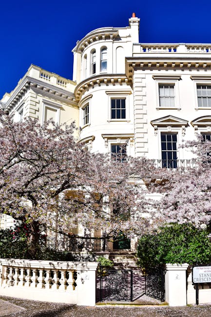 A large, elegant white Victorian-style building with detailed architectural features, including rounded bay windows and ornate cornices, is set against a bright blue sky. In the foreground, flowering trees with pale pink blossoms partially conceal the lower portion of the building, including a white balustrade and a decorative gate. The building appears to be situated on a slightly elevated position, with steps leading up from a paved pavement area. The scene is well-lit by natural sunlight, highlighting the clean and smooth finish of the building's exterior, which contrasts with the soft, textured blossoms of the trees. The environment suggests an urban residential setting, with the focus on the architectural detail and the seasonal flowering, relevant to inspections or maintenance of historic or private properties involved in waste and rubbish management services, as provided by Rubbish Removal Notting Hill.