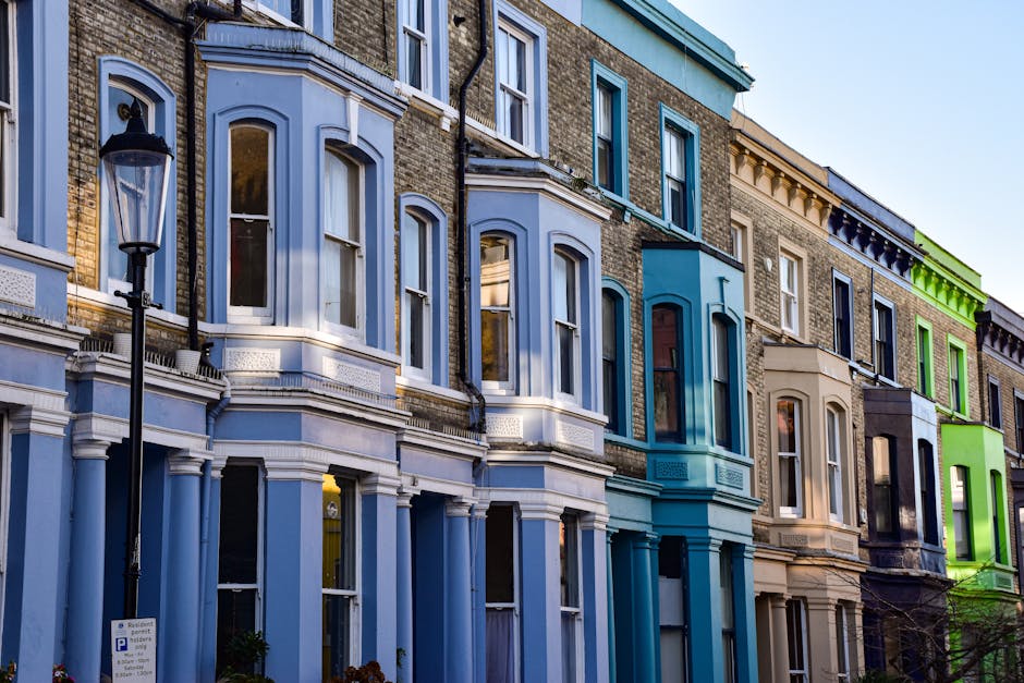 A row of Victorian-style terraced houses in a city neighborhood, each with distinctive features including large sash windows, decorative cornices, and bay windows. The buildings are constructed with brickwork, some painted in pastel shades such as light blue, cream, and green, with finishes ranging from unpainted brick to smooth painted surfaces. The facades display a variety of textures, with the painted sections appearing smooth and glossy, while the brickwork is rougher. The windows are framed with white trims, and some have white curtains visible inside. In the foreground, there is a black lamppost with a traditional lantern-style light fixture, positioned on the pavement adjacent to the houses. The scene is illuminated by natural daylight, with a clear sky visible in the background. The street environment suggests an urban residential area where private property maintenance, including rubbish collection and waste removal, may be managed through independent service providers like Rubbish Removal Notting Hill, supporting alternative waste handling arrangements for townhouses.