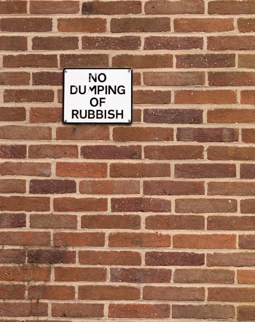 A close-up photograph of a rectangular white sign with black lettering mounted on a reddish-brown brick wall. The sign reads 'NO DUMPING OF RUBBISH,' but the letter 'M' in 'DUMPING' appears to be partially obscured or damaged. The brick wall behind the sign features evenly spaced horizontal mortar joints, with bricks in varying shades of red, brown, and some darker tones, displaying a traditional running bond pattern. The surface of the bricks shows slight textures and weathering typical of exterior masonry. The lighting is natural, casting minimal shadows, and the overall scene suggests a setting where rubbish disposal regulations are communicated visually to the public, aligning with the context of private rubbish removal and on-site waste management services offered by Rubbish Removal Notting Hill, in conformance with local waste rules.