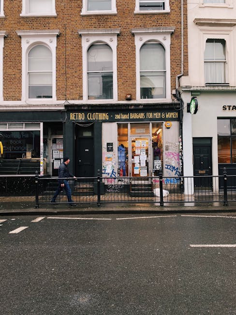 The image shows a street-level view of a small retail shop named 'Retro Clothing' with a black storefront and yellow lettering advertising secondhand bargains for men and women. The shop's large front window displays various items and notices, with a black door slightly left of centre. The building above features traditional brickwork with three arched sash windows, each framed by decorative white trim. To the right of the shop, part of another storefront with a white facade and a small sign is visible. In front of the shop, a pedestrian in dark clothing and a beanie walks along the pavement, which is separated from the road by a black metal railing. The road surface appears wet, likely from recent rain, and the muted daylight creates a subdued atmosphere. This scene visually aligns with private or independent waste management activities typical of small retail premises, such as rubbish removal or on-site clearance services provided by companies like Rubbish Removal Notting Hill, especially considering the urban environment suited for domestic or commercial waste collection in a bustling high street area.
