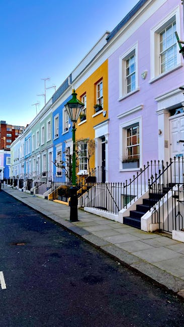 A row of Victorian terraced houses painted in pastel shades of blue, yellow, and lavender along a quiet urban street. Each property features white-framed sash windows, small front gardens with black iron railings, and steps leading up to front doors. A traditional black street lamp stands on the sidewalk in front of the yellow house, casting a subtle reflection on the pavement. The scene is illuminated by natural daylight, with clear blue skies overhead and a few television antennas visible atop the roofs. The street appears clean and well-maintained, representing typical residential surroundings where private rubbish collection services, such as those provided by Rubbish Removal Notting Hill, are often used for on-site clearance or alternative waste handling. The pavement runs smoothly alongside the houses, and the overall atmosphere is calm and orderly, emphasizing the neat urban environment that may occasionally require rubbish removal for property upkeep or decluttering purposes.