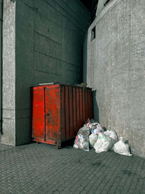 A large, rusted metal dumpster with a red exterior and vertical ridges, positioned in the corner between a dark grey building wall and a light grey concrete wall on a paved sidewalk. Several white plastic garbage bags filled with mixed waste materials are piled against the base of the dumpster, some leaning slightly, with recyclables or general waste visible through semi-transparent bags. The scene is outdoors, with subtle shadows indicating daylight, and the environment appears to be an urban setting, possibly in an alleyway or side street. The presence of the waste suggests alternative private rubbish disposal options, with Rubbish Removal Notting Hill offering on-site clearance services for such waste accumulation, supporting effective and immediate rubbish removal from commercial or residential sites.