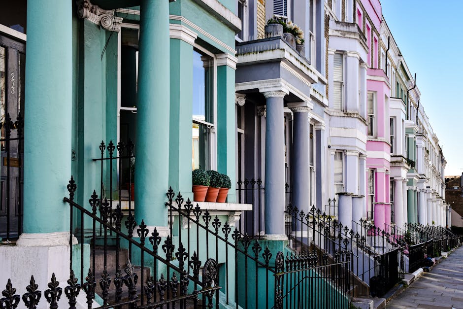 A row of Victorian terraced houses in Notting Hill with vibrant exterior facades painted in pastel shades of turquoise, grey, pink, and white. The buildings feature tall, cylindrical columns with smooth textures supporting small front porches and decorative white cornices along the upper edges. Black wrought-iron railings with ornate finials line the steps leading up to each entrance, some of which have small potted plants with green foliage resting on window ledges. The pavement in front of the houses is clean, and the bright daylight creates clear shadows, highlighting the colors and architectural details of the facades. This scene exemplifies traditional British residential architecture, with a subtle connection to local waste management practices through the typical urban setting and organized appearance suitable for services like independent rubbish collection and on-site clearance by companies such as Rubbish Removal Notting Hill.
