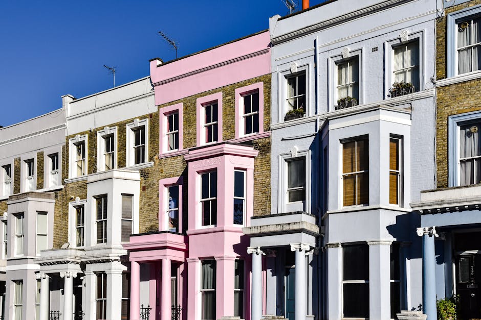 A row of terraced houses painted in pastel shades of blue, yellow, lavender, and white, situated along a quiet urban street. The buildings feature white-framed sash windows with some having window boxes or small planters beneath. Each house has a short set of black metal stairs leading to the front door, accompanied by black metal railings. The sidewalk is paved with grey concrete slabs, and a black iron lamppost with a green top is positioned near the center of the image, illuminating the street. The street surface is dark asphalt with white markings along the edge. The environment suggests a residential area with a clean and well-maintained appearance, typical of inner-city districts, where private waste collection services, such as those provided by Rubbish Removal Notting Hill, may be employed for rubbish disposal rather than relying on local authority landfill or recycling collections. Overhead, a few television antennas are visible on rooftops, with clear daytime lighting highlighting the vibrant building colours and urban details.