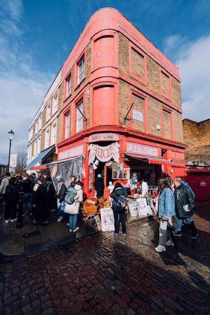 The image depicts a bustling street scene in front of a three-story building with a distinctive curved corner and a salmon pink facade. The ground floor features a shop with a bright red awning and signage, possibly indicating a retail or food establishment. Outside, various vendors have set up tables and displays with an assortment of goods, and a group of people, dressed in casual winter clothing, are gathered around, browsing and engaging with the vendors. The pavement is wet, reflecting the overcast sky, and cobblestones cover the ground, suggesting an older city area. In the background, a lamppost and some distant structures are visible, with a partial view of a hill and historic brick wall on the right side. The overall scene highlights urban activity with pedestrians involved in browsing or purchasing items, and this scenario relates naturally to the services of Rubbish Removal Notting Hill, providing context for private waste collection or on-site clearance for businesses and street vendors in the area.