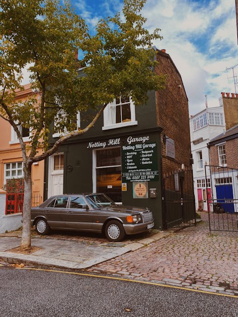 A two-storey brick building with a dark green facade and large white signage reading 'Notting Hill Garage' and 'Notting Hill Garage' in smaller font, situated on a cobbled street in Notting Hill. In front, a silver vintage car is parked partly on the pavement and partly on the driveway. To the left of the building, a leafy tree with yellow-green leaves extends over the car, providing partial shade. The building features a window on the upper floor with white frames, and the ground floor has a wide window and entrance door, both partially visible. Next to the building, a small iron gate and fence surround a yard or parking area. The environment appears quiet and residential, with neighboring houses visible in the background, and the sky above is partly cloudy with patches of blue. The scene reflects elements of private waste handling and on-site clearance, typical of commercial properties in the area, with the presence of the vehicle and signage indicative of independent collection and alternative waste management approaches by local services.